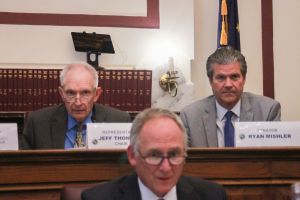 Jeff Thompson and Ryan Mishler sit at a long desk. Thompson is a White man with white hair. He is wearing glasses and a dark suit, blue shirt and gold patterned tie. Mishler is a White man with dark hair. He is wearing a light gray suit.