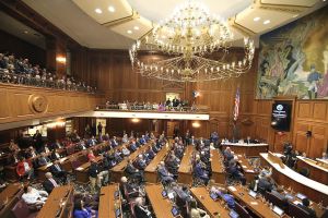Eric Holcomb stands in front of a microphone in the Indiana House. Holcomb is a White man with white and gray hair and beard. He is wearing a blue suit.
