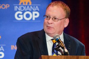 Mike Braun speaks at a lectern on a stage. Behind him are an American flag and a backdrop with his campaign logo. Braun is a White man, balding, with gray hair. He is wearing glasses and a suit.