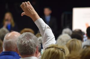 Andrew Ireland stands on the House floor looking at Matt Pierce, who is in the foreground, standing in profile and raising his hand while he speaks. Ireland is a White man with dark hair, wearing a suit and tie. Pierce is a White man with gray hair